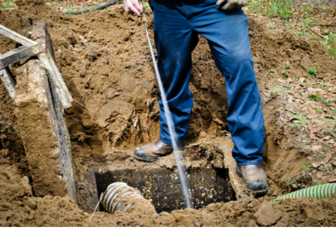 Man in blue jeans, boots, standing in excavated soil, inspecting septic tank.