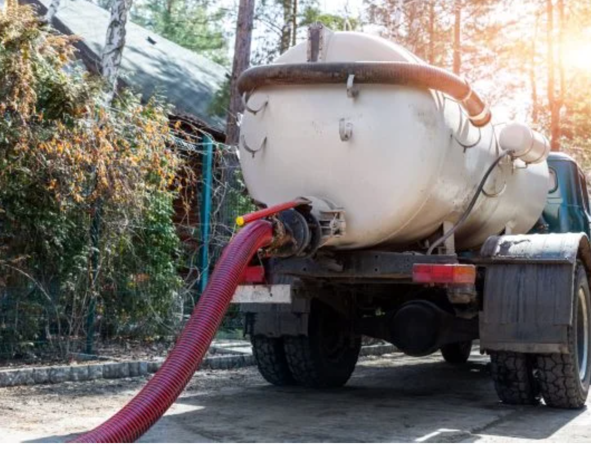 Septic tank truck discharging waste. Red hose extends from white tank. Outdoor setting.