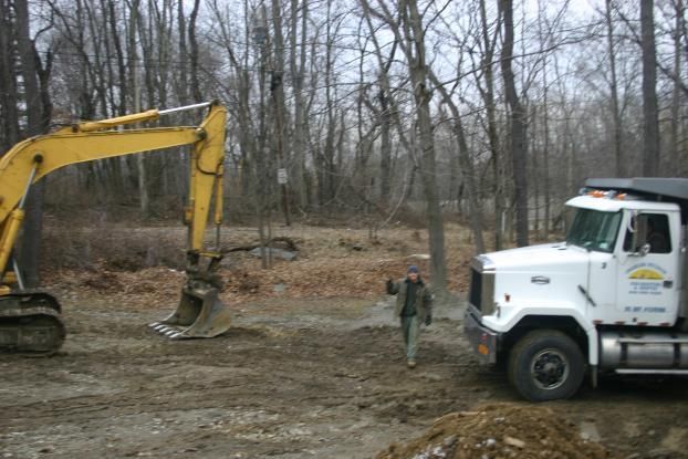 Excavator and dump truck at a construction site, man walking towards truck, trees in background.
