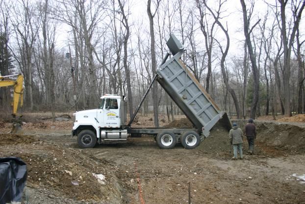 Dump truck dumping material in a construction site with two people watching, surrounded by trees.