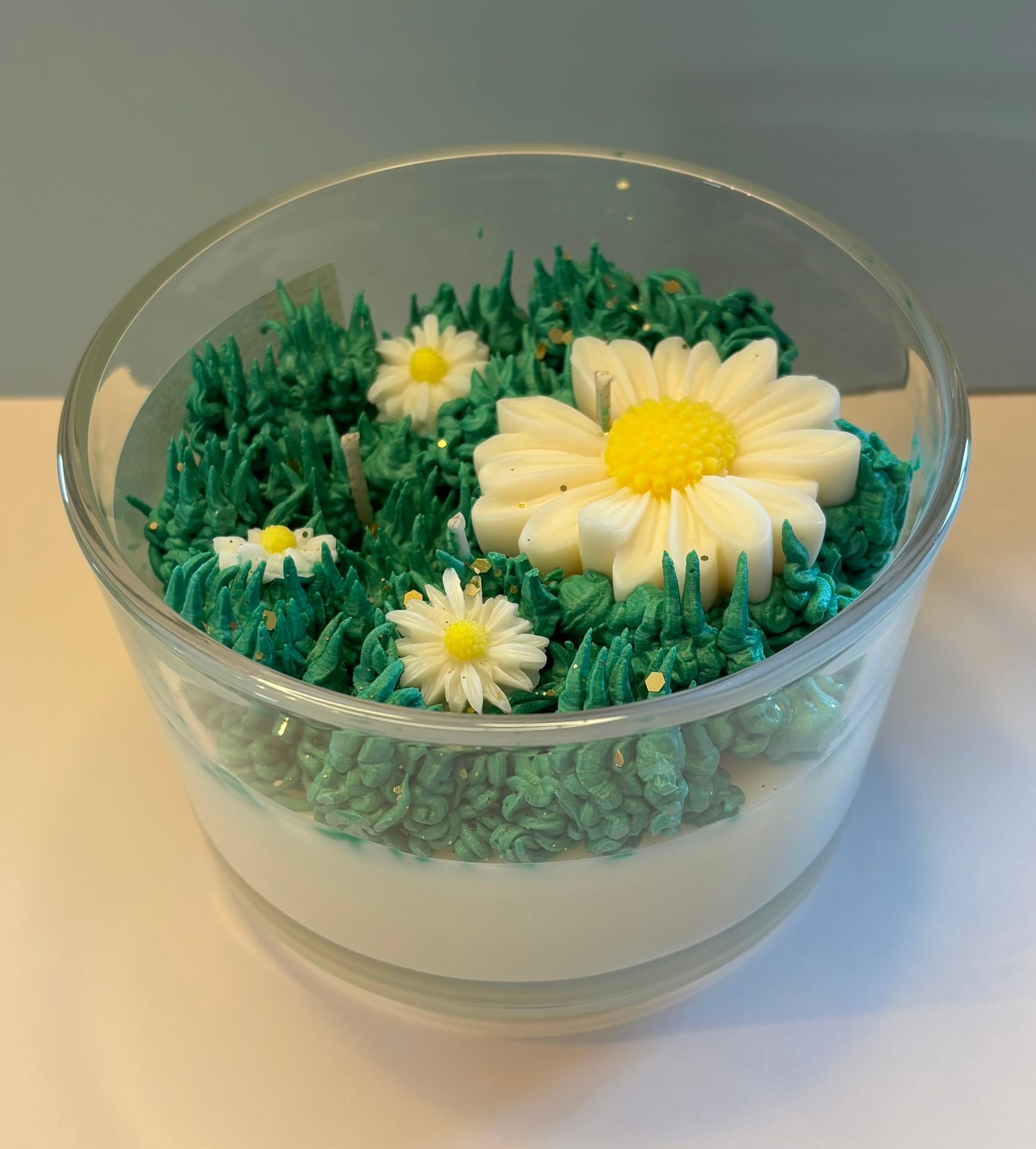 Een kaars in een doorzichtige glazen kom, versierd met groen wasgras en diverse witte en gele madeliefjes.