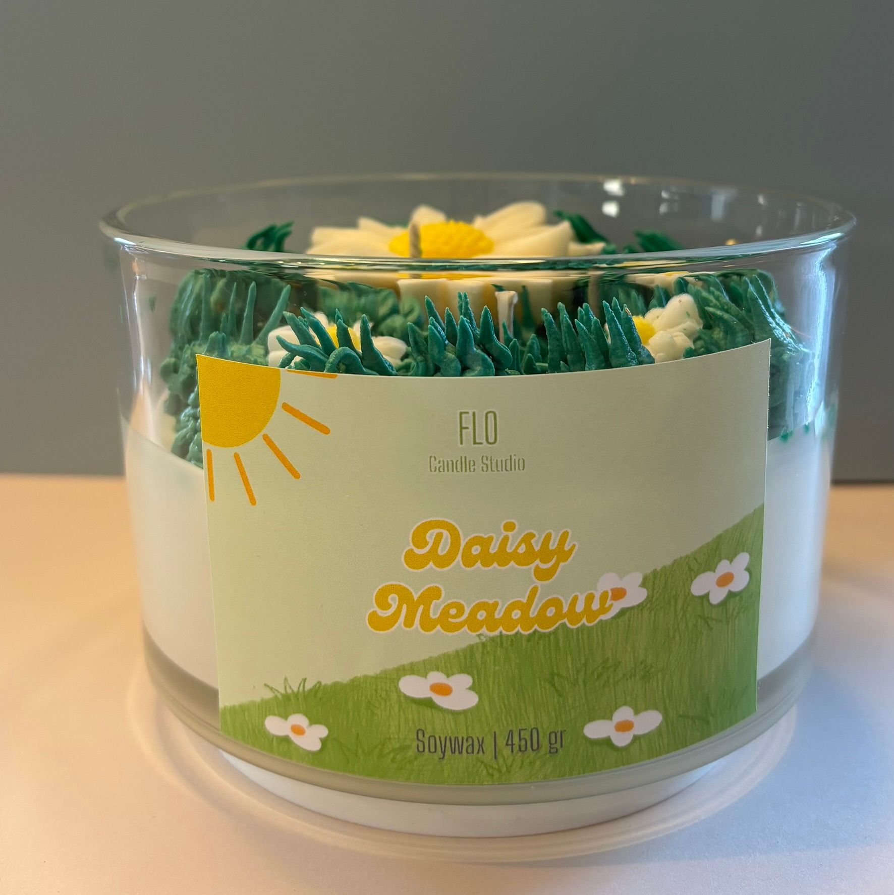 Een kaars in een doorzichtige glazen kom, versierd met groen wasgras en diverse witte en gele madeliefjes.