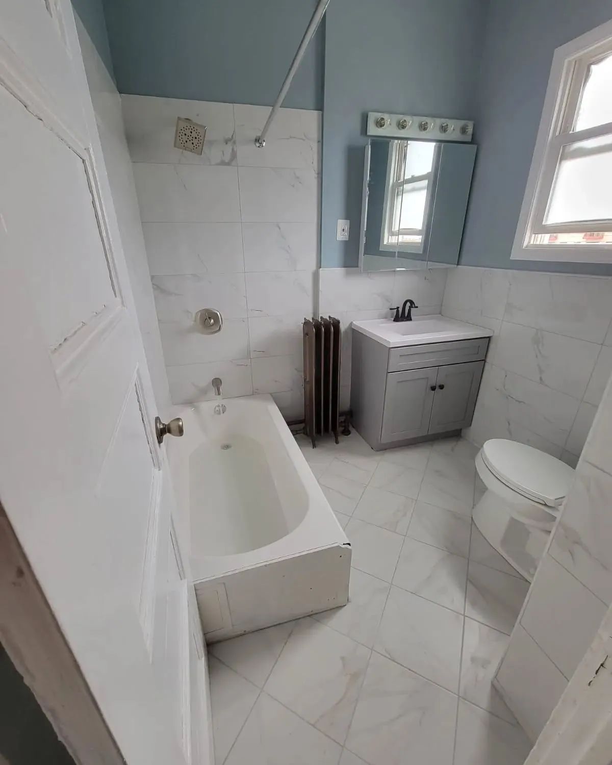 Bathroom with white tile, tub, sink, and toilet. Blue walls and window, with a mirror above the sink.