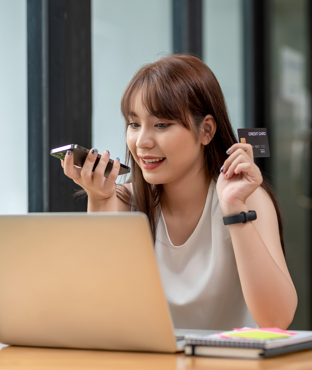A woman is sitting in front of a laptop computer holding a credit card and a cell phone.