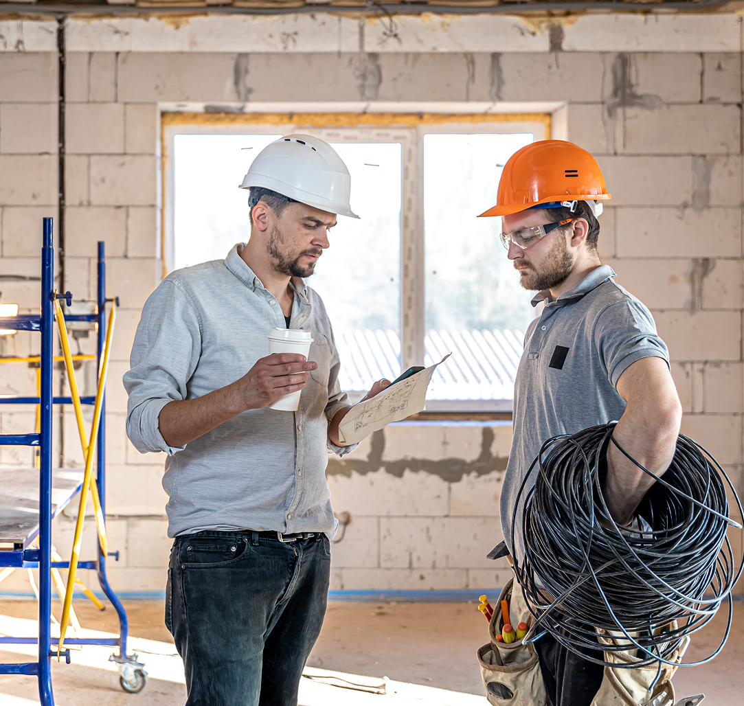 Two construction workers are standing in a room looking at a clipboard.