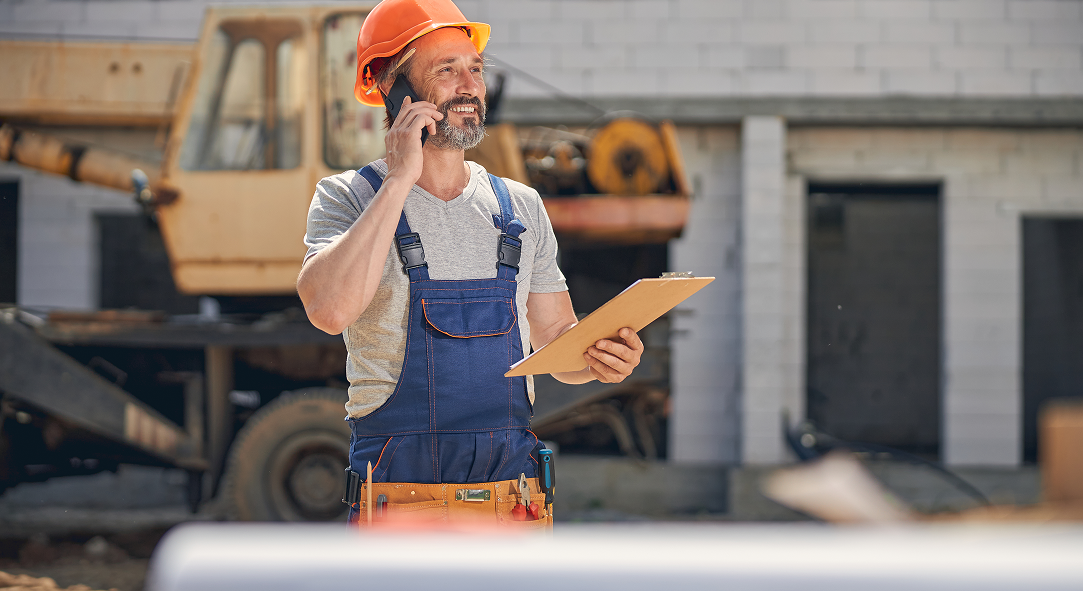 A construction worker is talking on a cell phone while holding a clipboard.