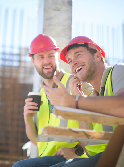 Two construction workers are sitting at a table drinking coffee and laughing.