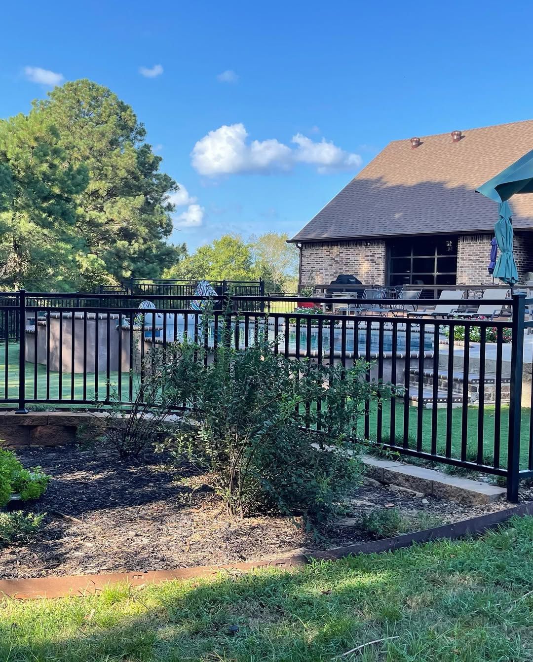 Black metal fence surrounds a backyard with a house, bushes, and a blue sky.