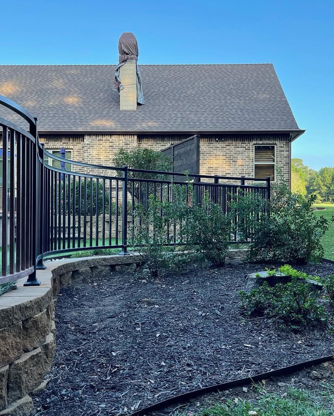 Black metal fence surrounds a garden bed in front of a brick house with a dark roof.