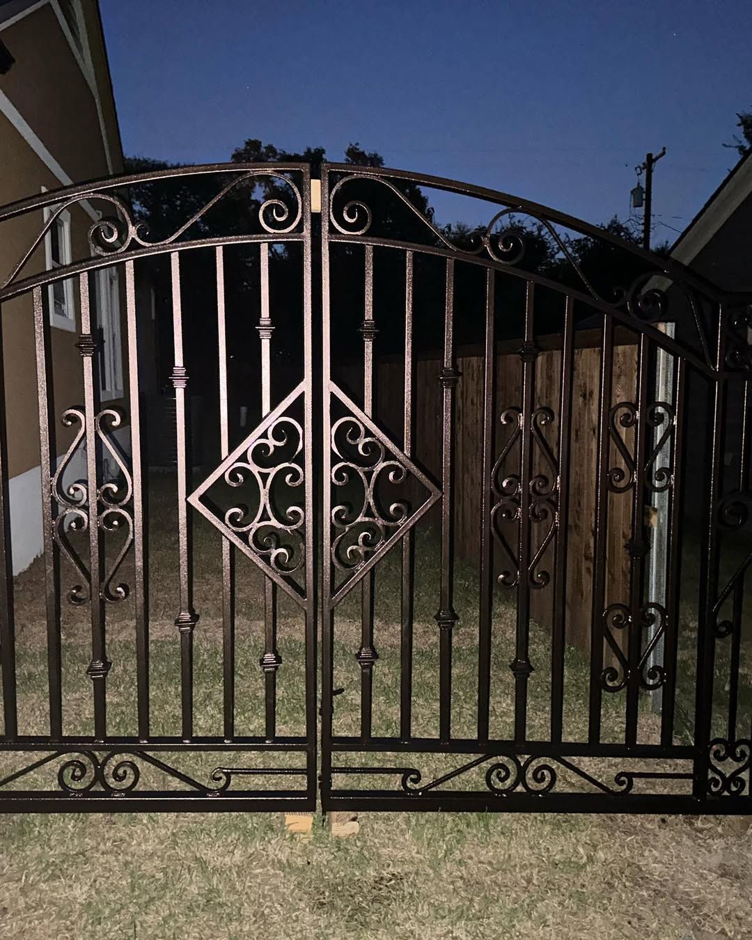 Ornate, dark metal double gate set in a grassy yard, with decorative scrollwork, against a wooden fence.