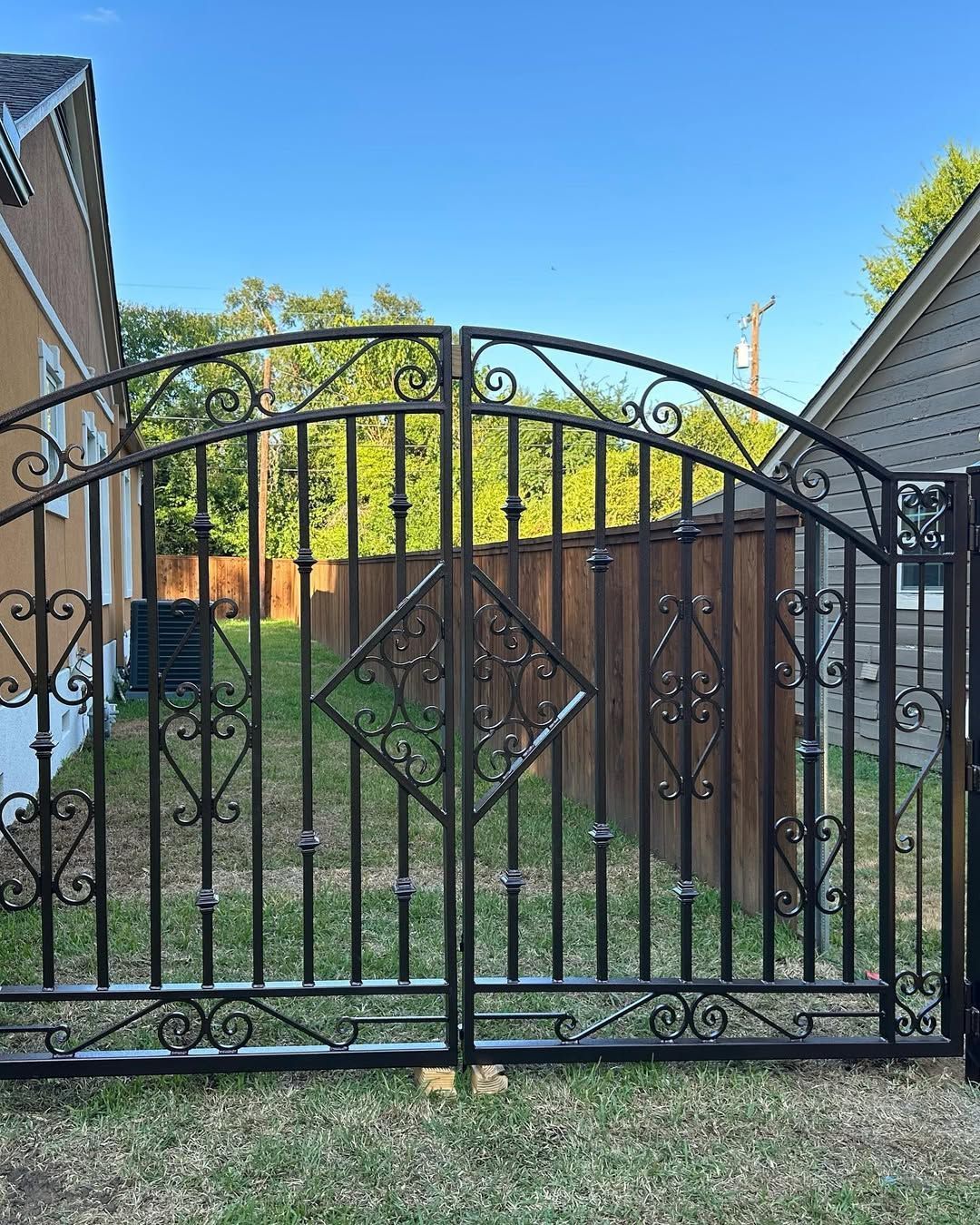 Black wrought iron double gate with ornamental details, in front of a wooden fence and yard.