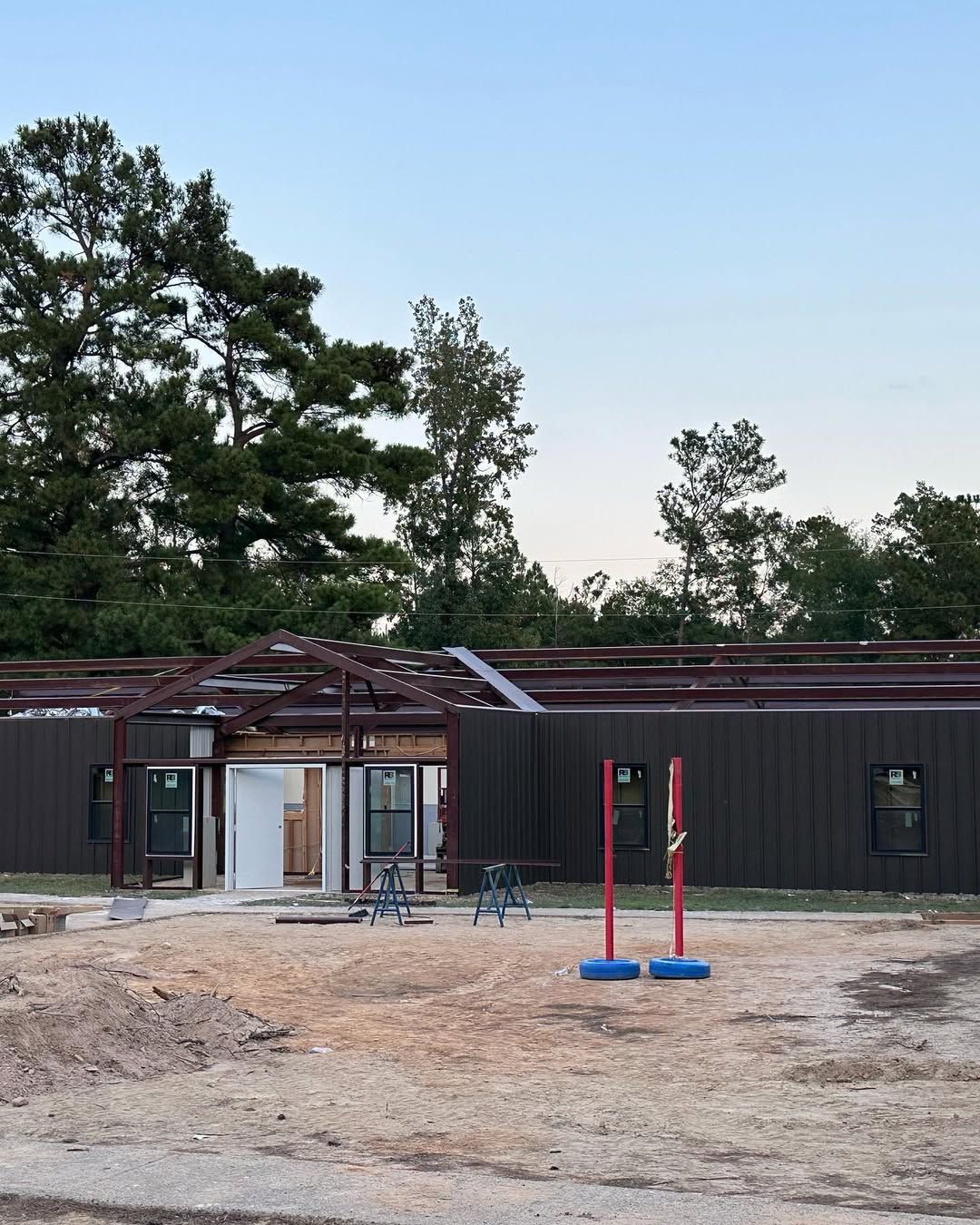 Construction site with partially built metal building, surrounding dirt, and trees in the background.