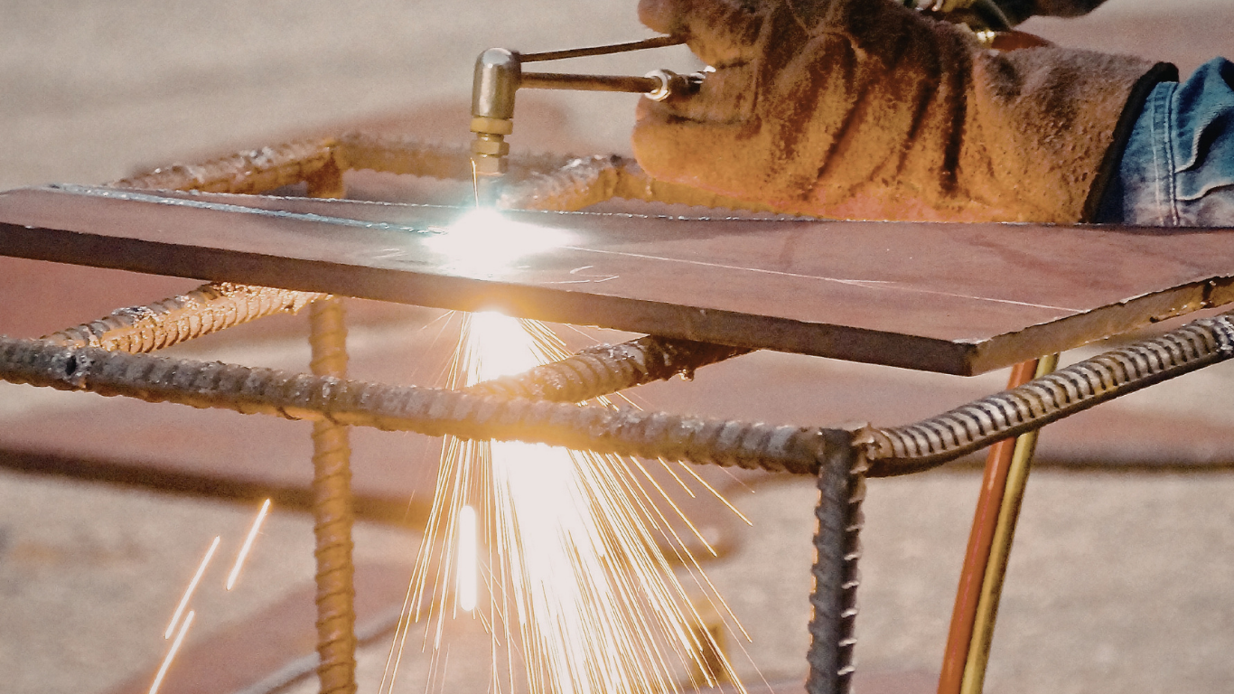 A person in work gloves uses a torch to cut through a metal sheet supported by rebar.