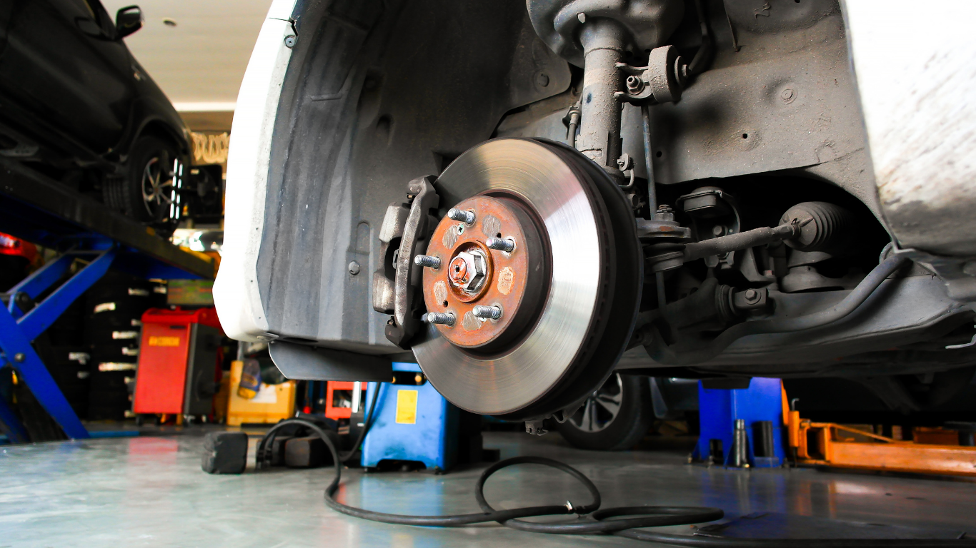 Car's brake disc and caliper are visible, wheels removed, in a mechanic's shop setting.