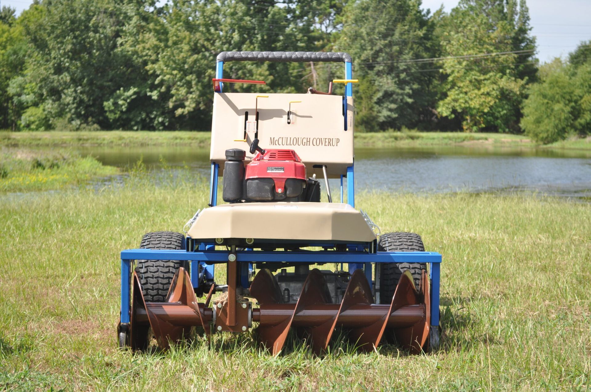 A beige and blue pond weed harvester in a grassy area next to a pond.