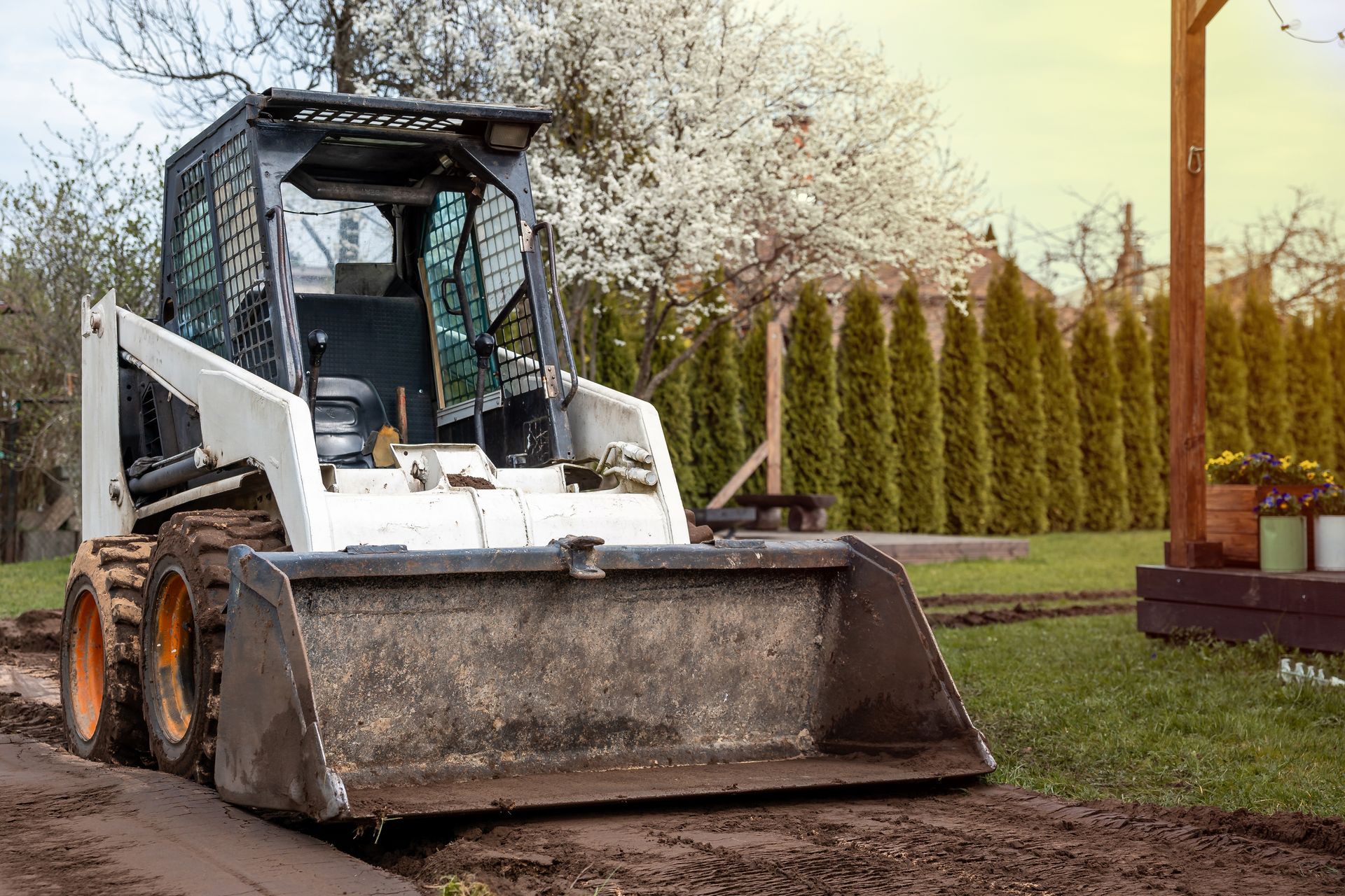 White skid steer loader on muddy ground near a grassy yard and wooden posts.