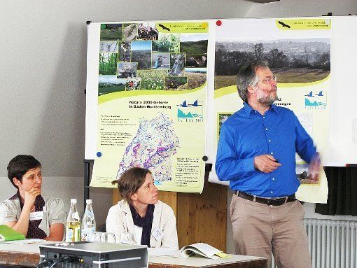 Ein Mann hält einen Vortrag auf einer Konferenz. Zwei Frauen sitzen an einem Tisch. Im Hintergrund hängen Plakate.