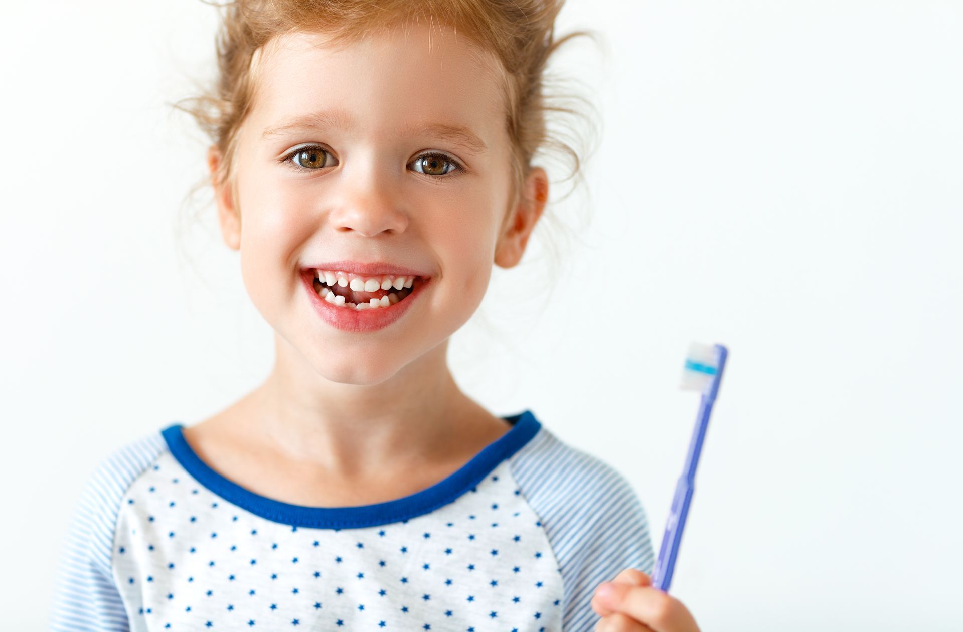 Smiling young girl with missing front tooth, holding a toothbrush against a white background.