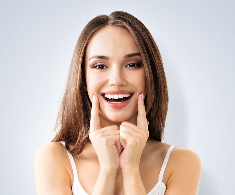 Femme souriante pointant ses joues. Elle a de longs cheveux bruns, porte un haut blanc et affiche un large sourire.