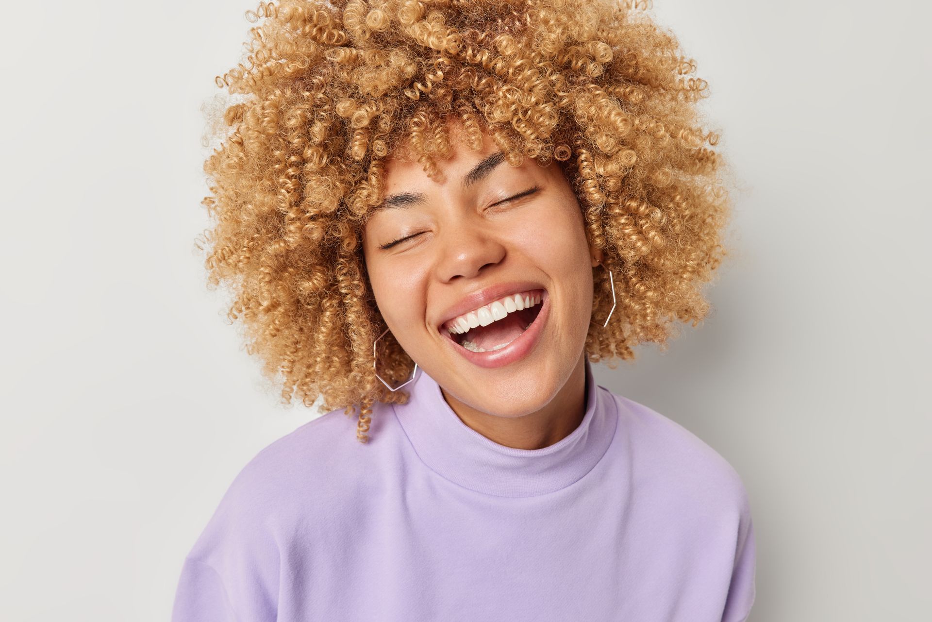 Une femme aux cheveux bouclés rit les yeux fermés et porte un pull violet.