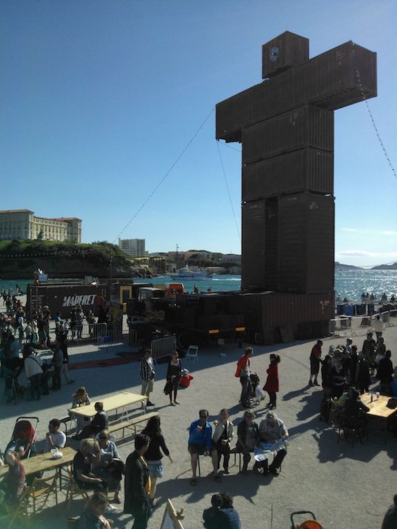 A human like sculpture out of containers at the seashore with a group of people sitting on benches around it.