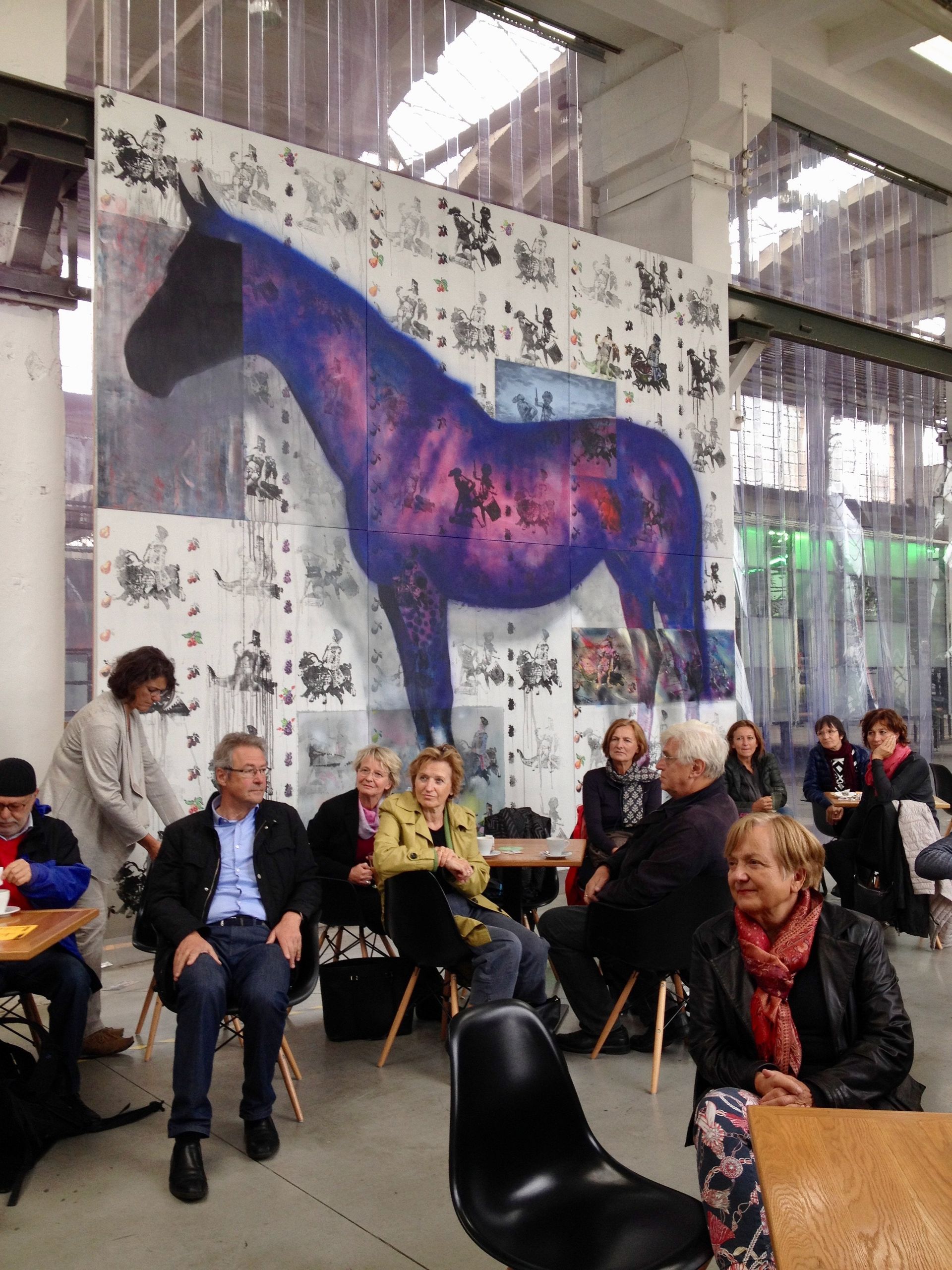 A group of people in a room, listening to a conference, in the background a huge poster showing a violet horse.