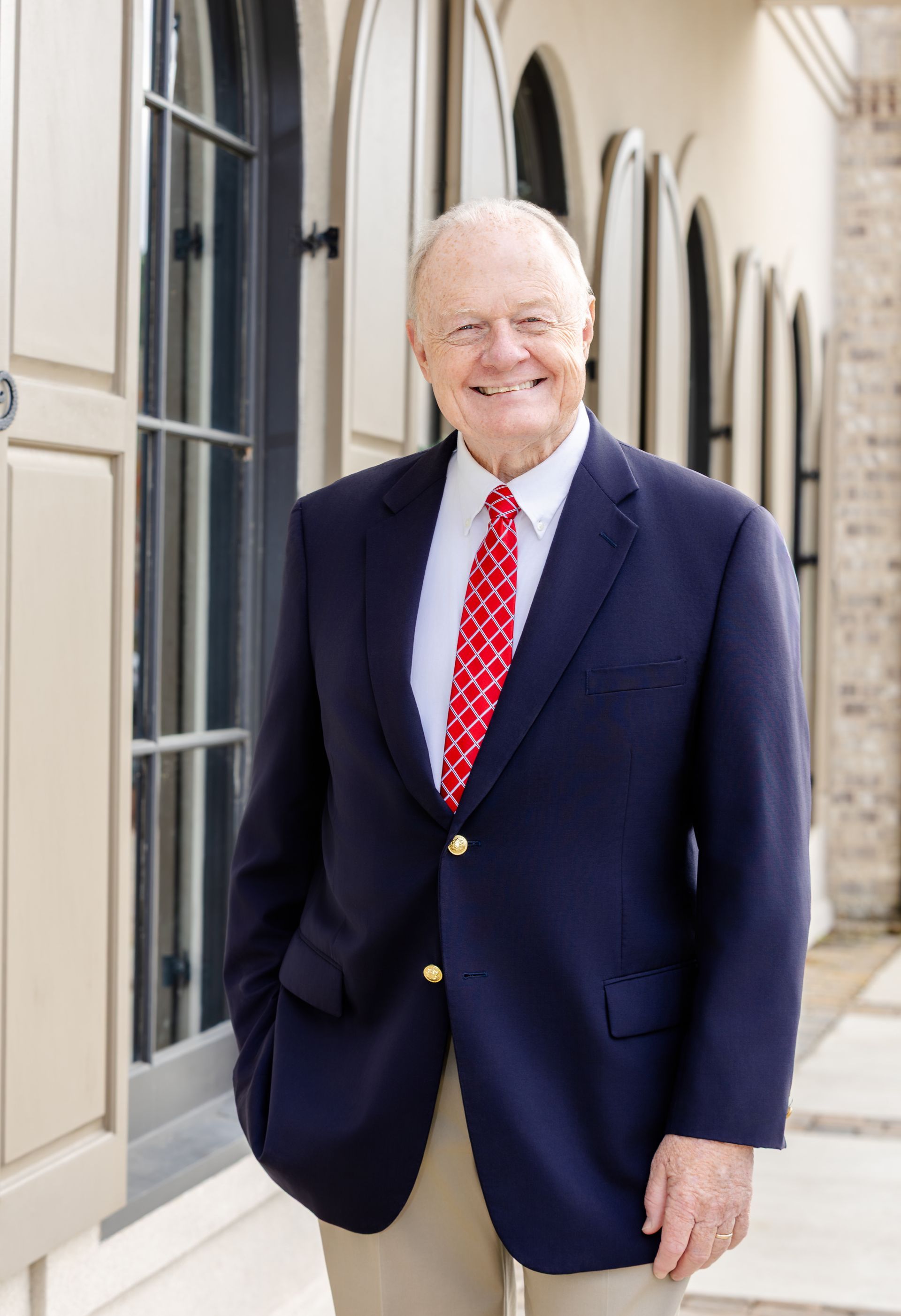 A smiling person in a blue blazer and red patterned tie standing outdoors near arched windows.