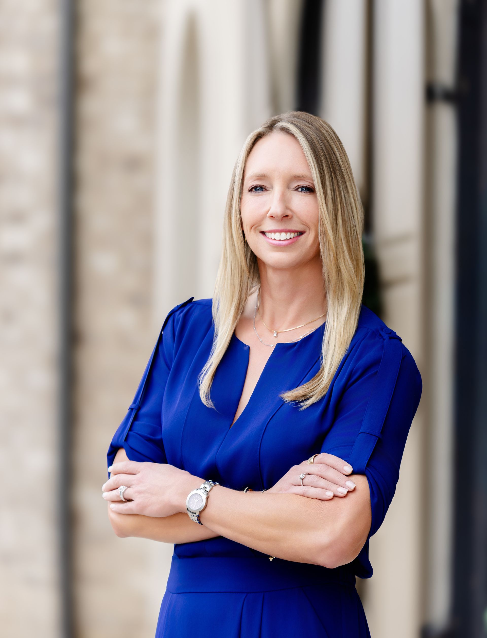 A professional woman with blonde hair, wearing a royal blue dress, smiling with arms crossed in front of a building.
