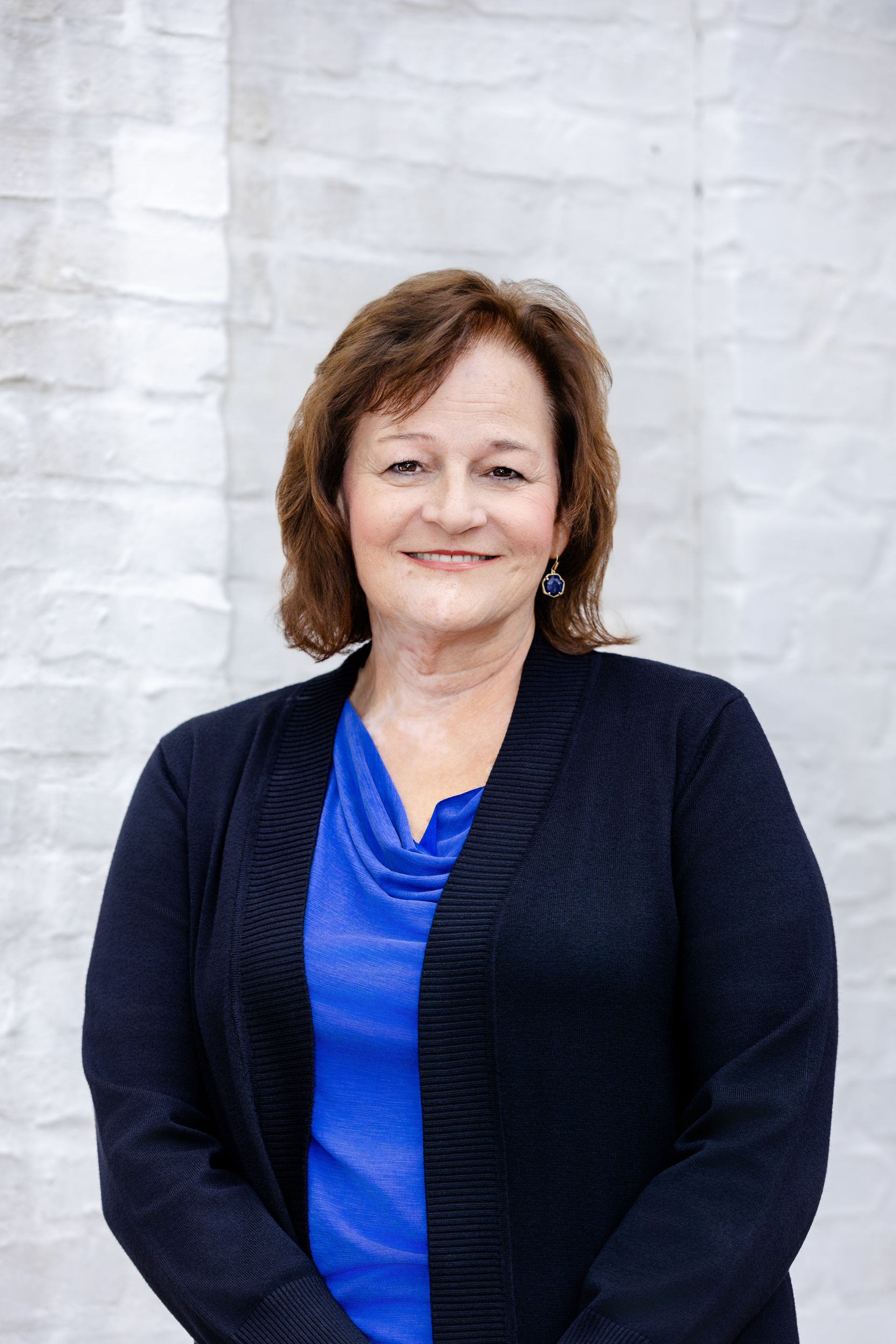 A smiling professional person wearing a blue shirt and dark cardigan against a white brick wall.