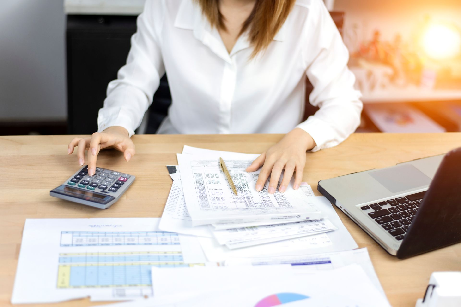 Woman in white shirt working at desk with calculator, documents, and laptop.