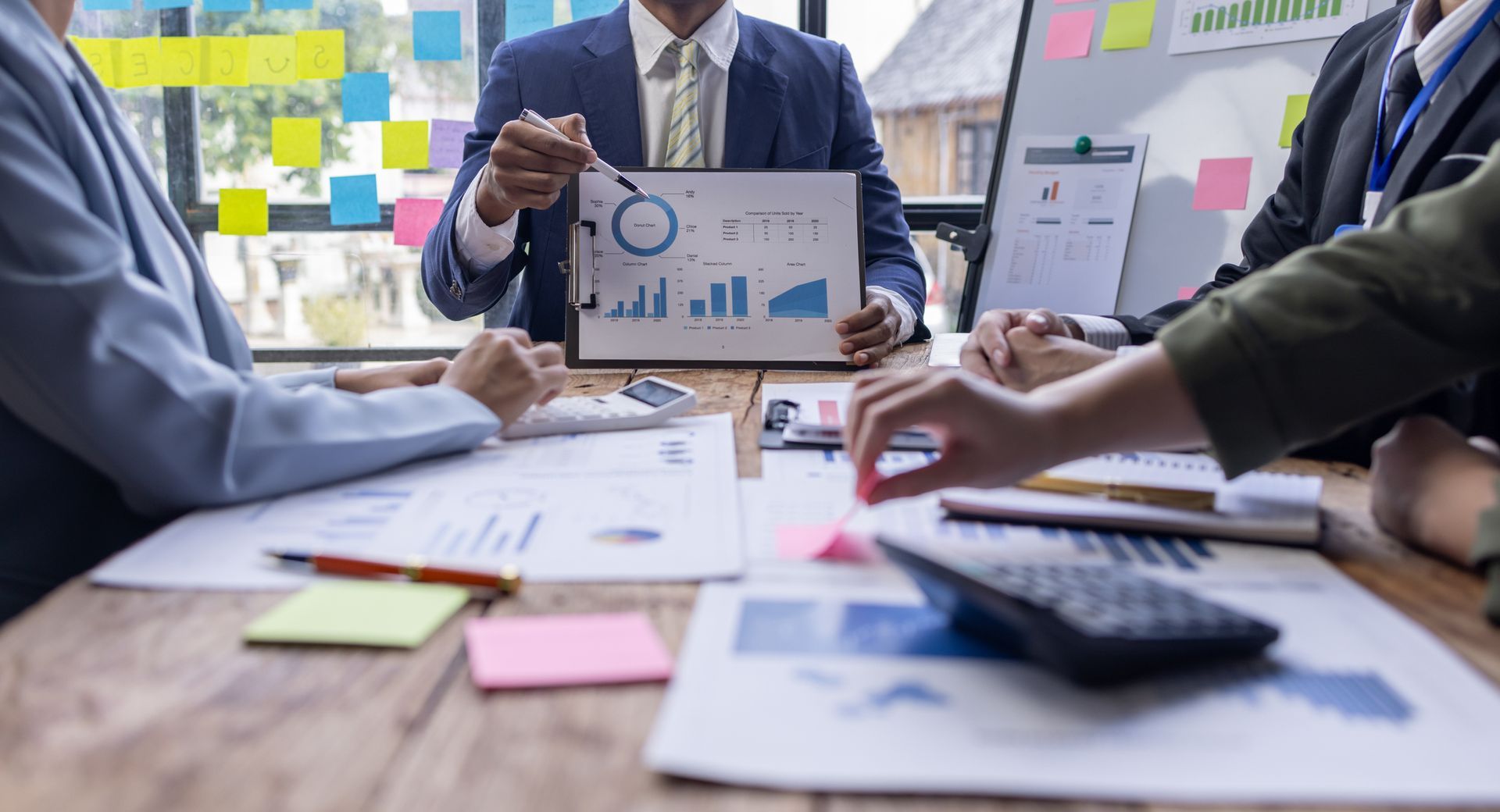 Business meeting with charts, documents, and calculator on a wooden table; a person presents data.