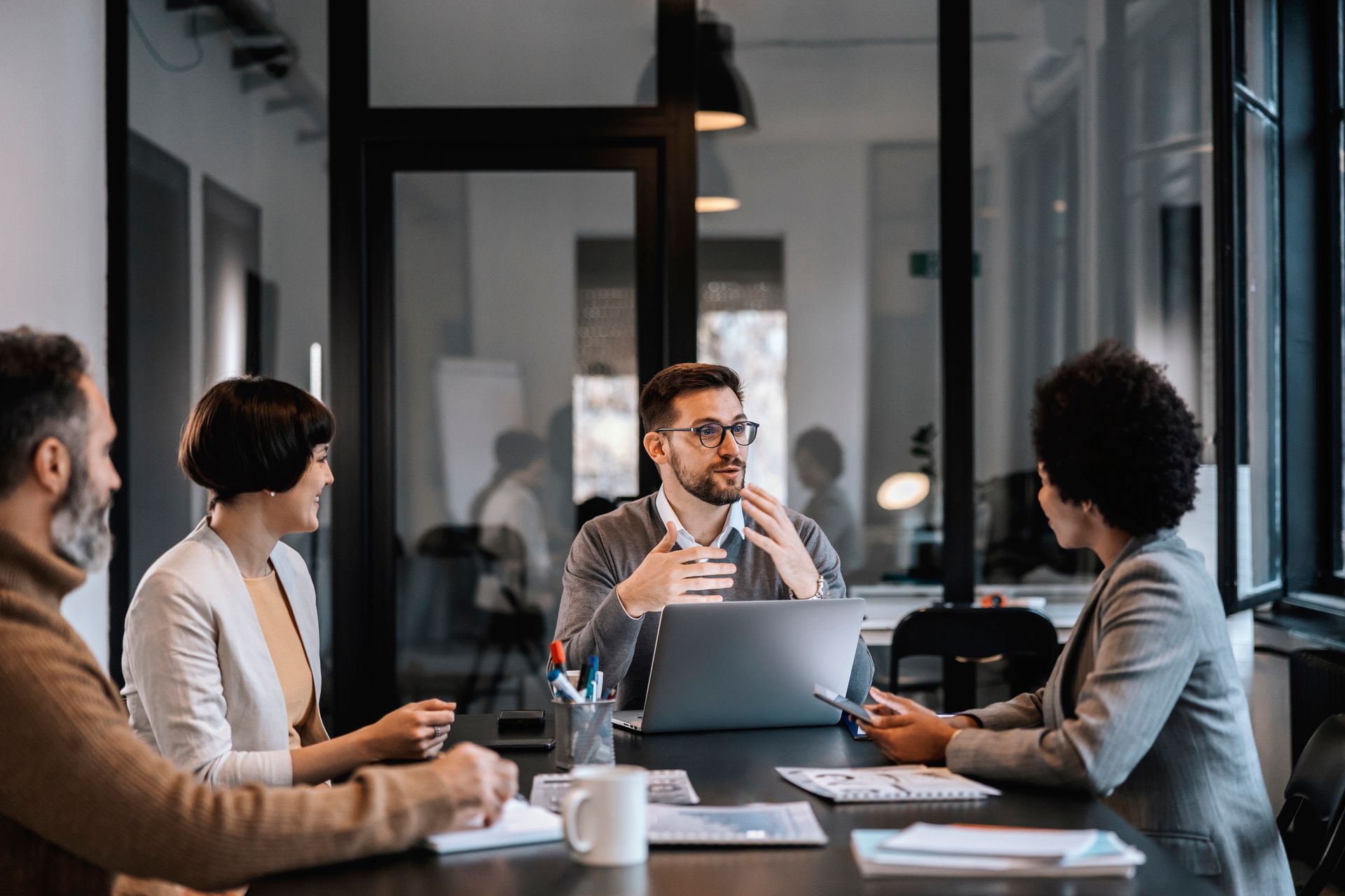 Four business professionals at a conference table, collaborating with a laptop, documents, and thoughtful expressions in an office setting.