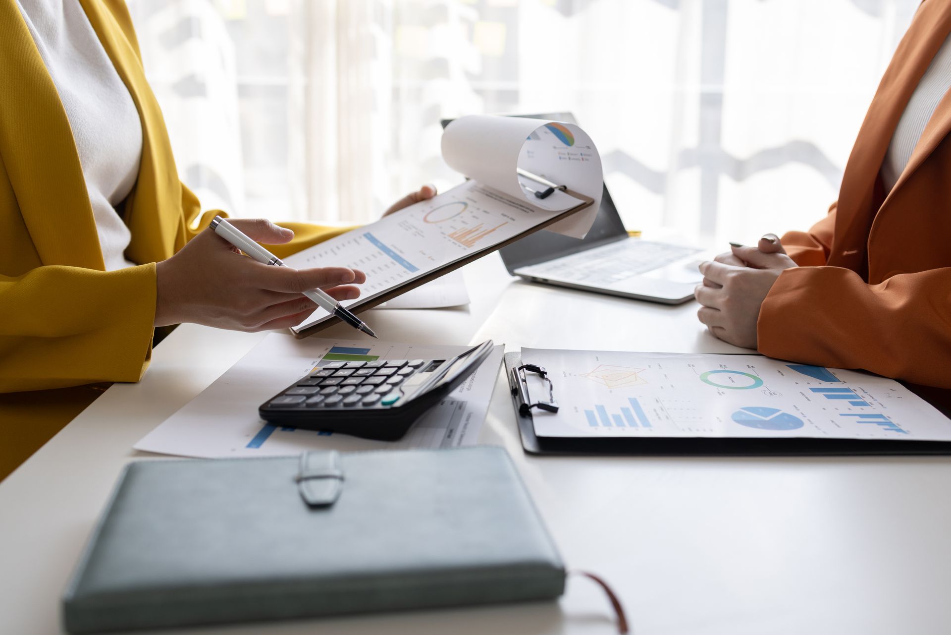 Two women in blazers review financial documents at a desk with a calculator and laptop.