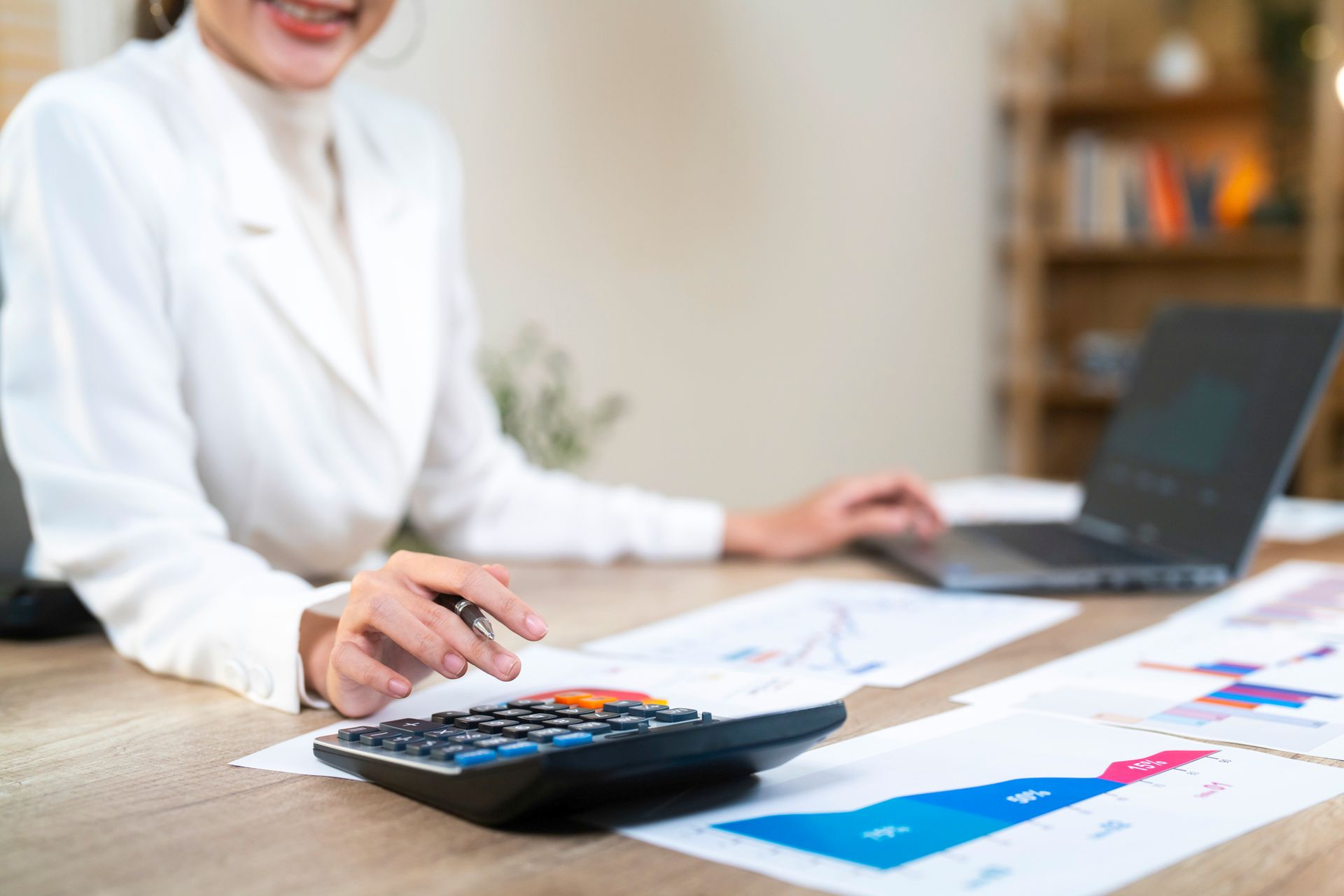 Woman in white jacket smiles while using a calculator, with charts and laptop on desk.