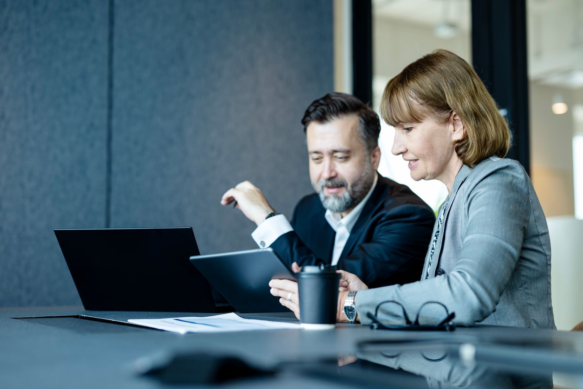 Man and woman reviewing documents on a tablet and laptop at a table in an office setting.