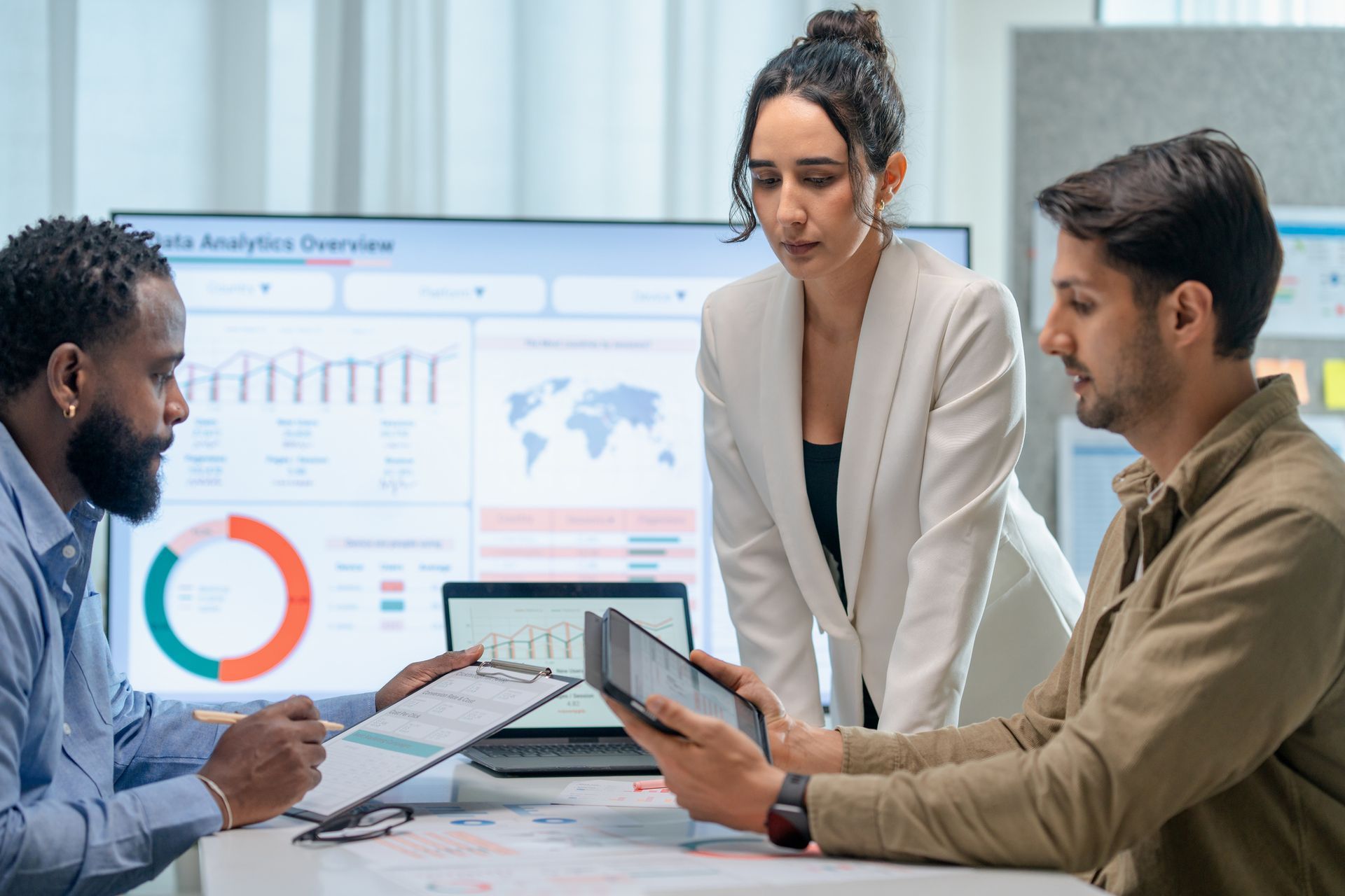 Three people in a meeting, looking at a tablet and papers, with a data analytics overview on the screen.