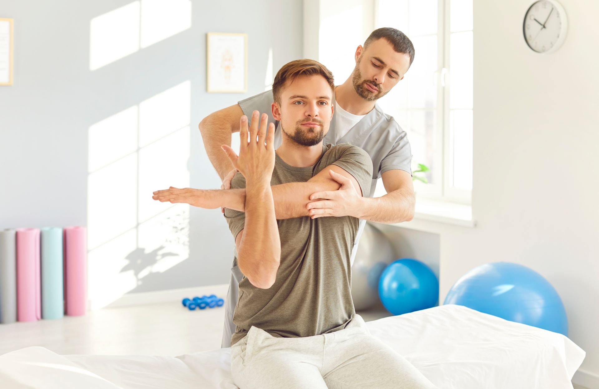 A male physical therapist helping a man stretch his shoulder in a bright therapy room.