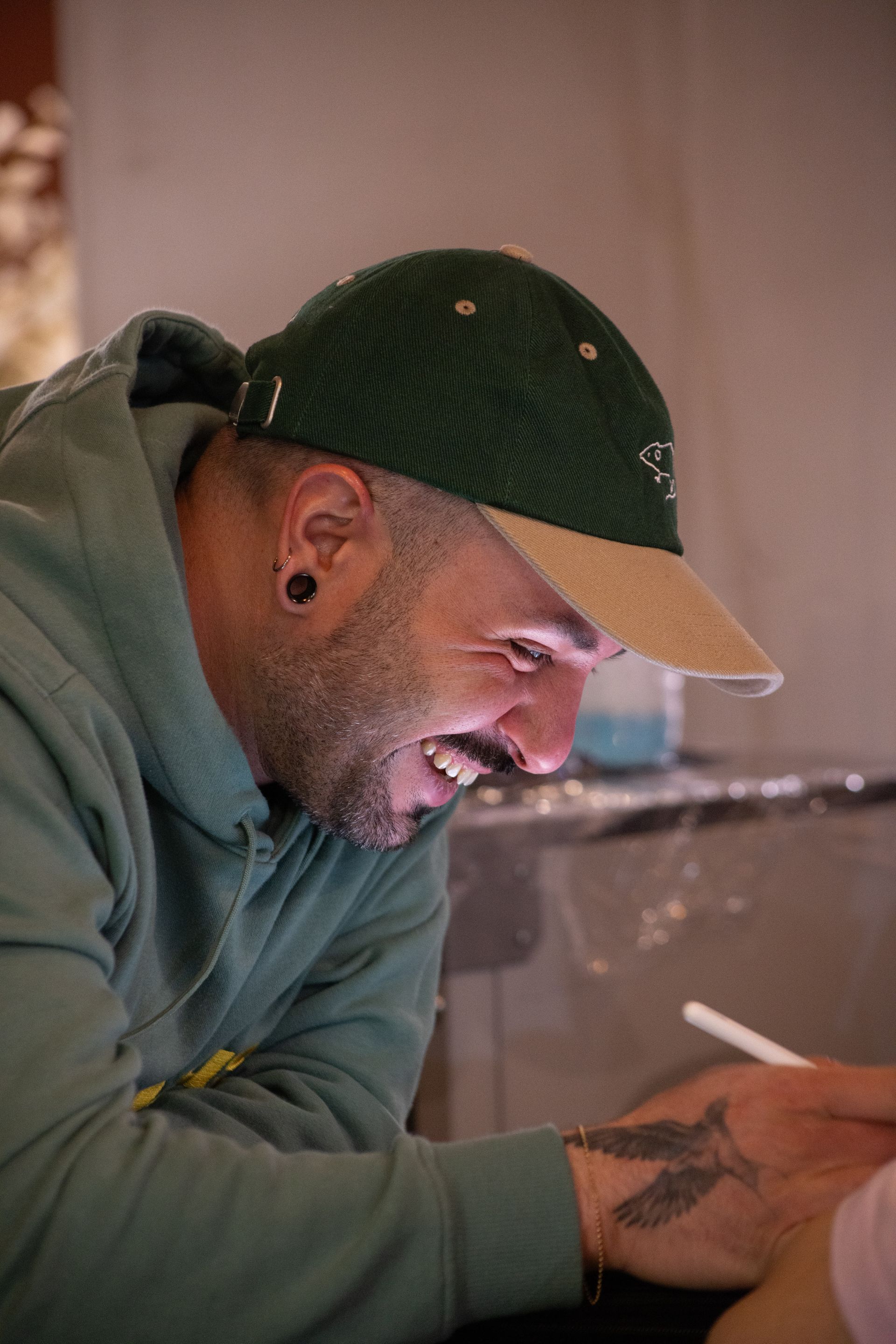 A person in a green hoodie and baseball cap smiling while working on a tattoo on a client's arm.