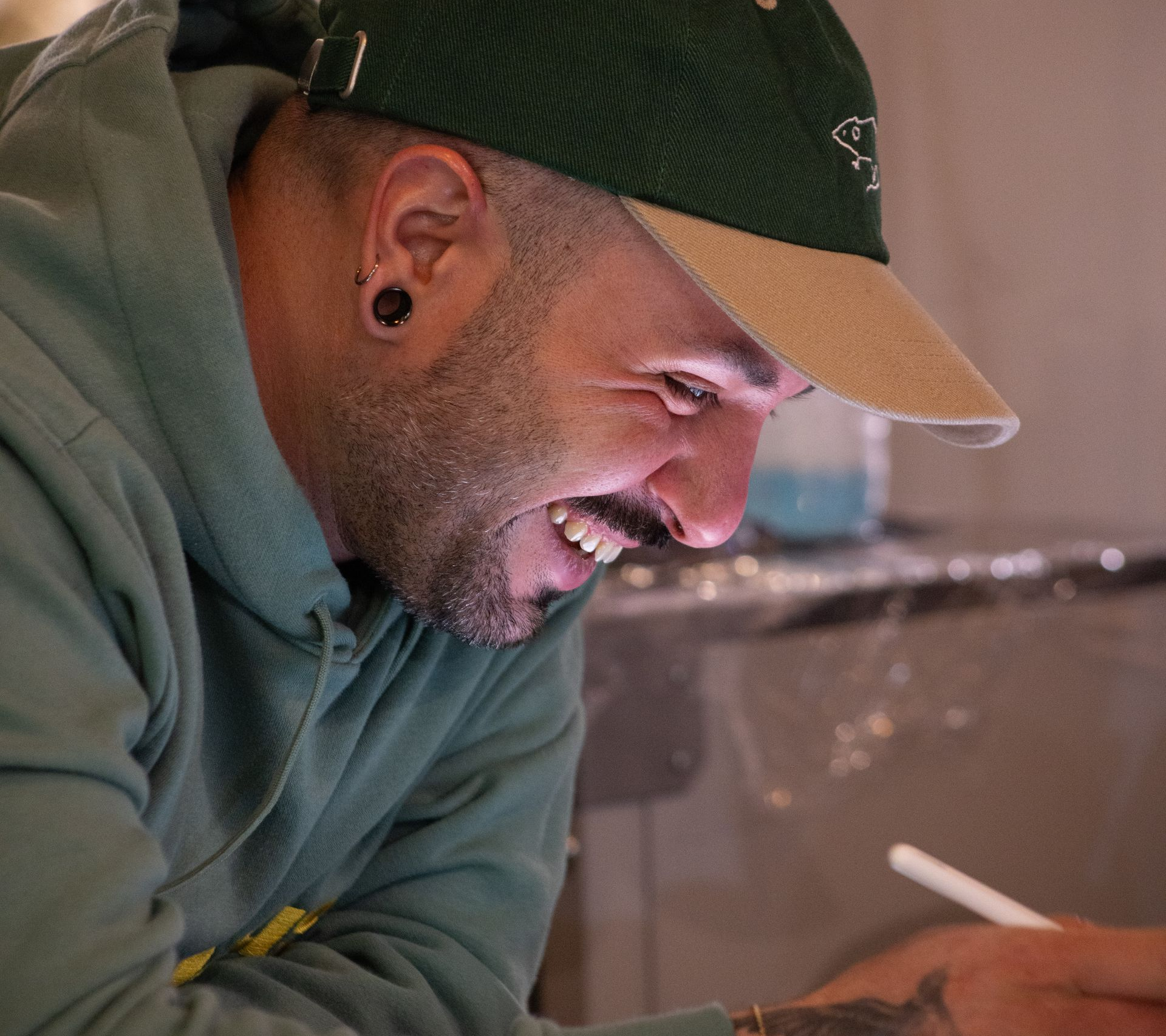A smiling person in a green hoodie and green baseball cap with a tan brim, looking down and working on a task.