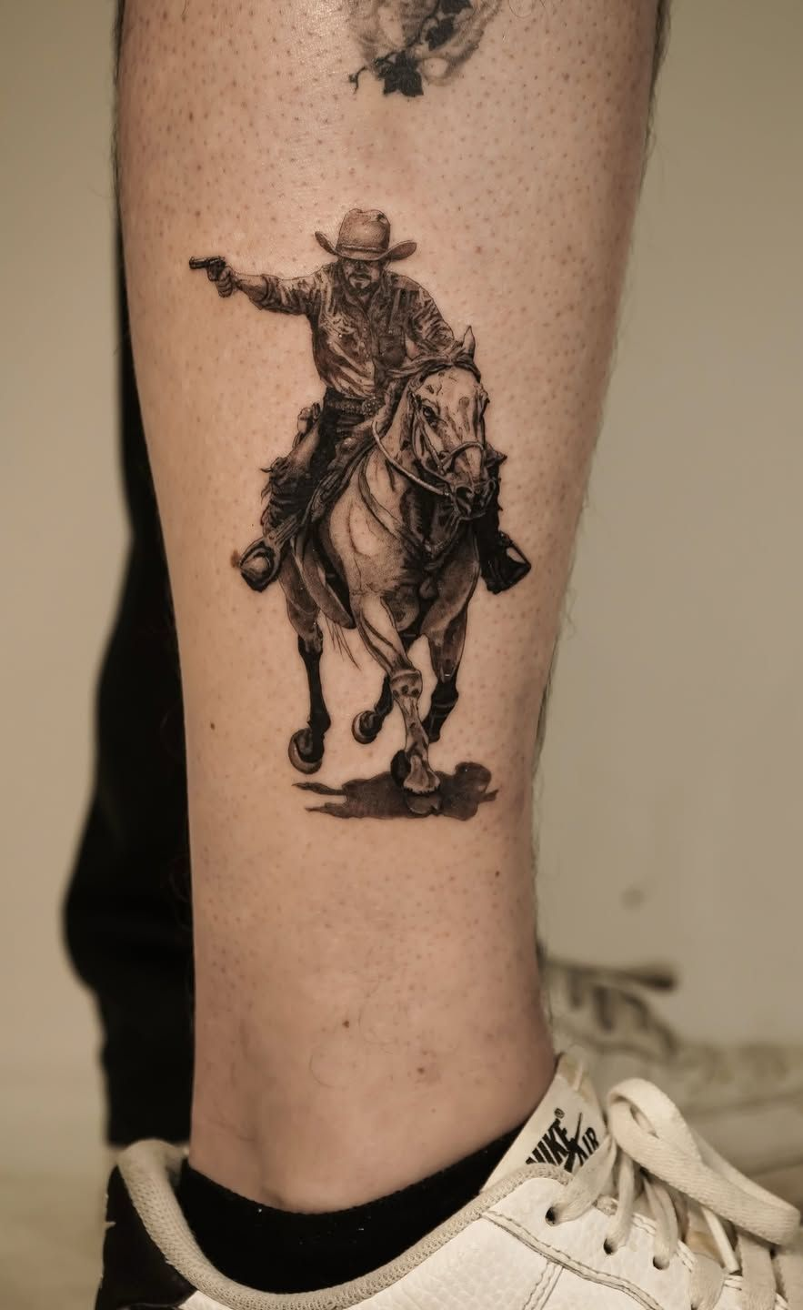 A black-and-grey tattoo on a person's leg depicting a cowboy riding a horse while pointing a handgun.