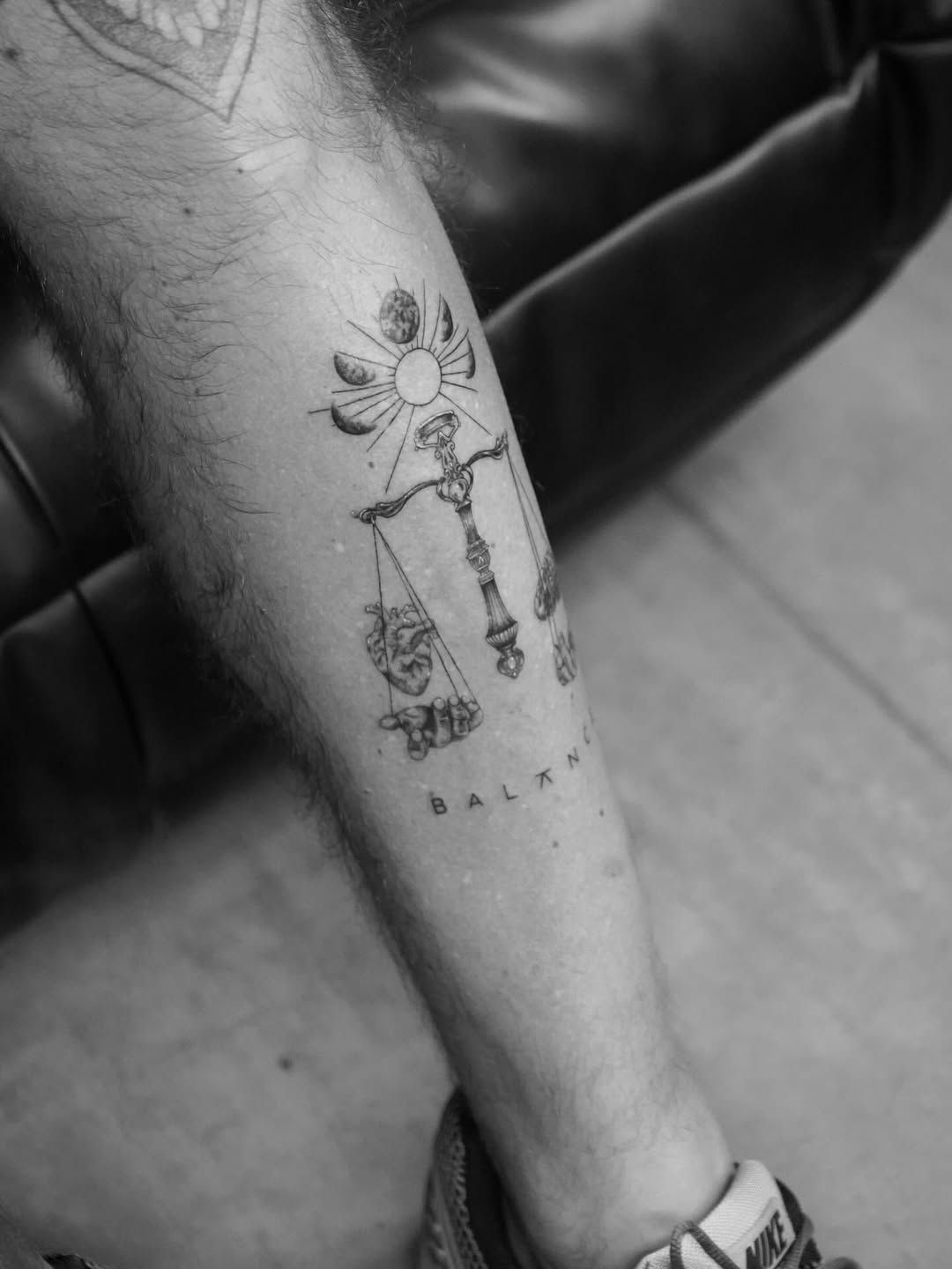 A black-and-white tattoo on a lower leg featuring a scale of justice, a sun with lunar phases, and the word 
