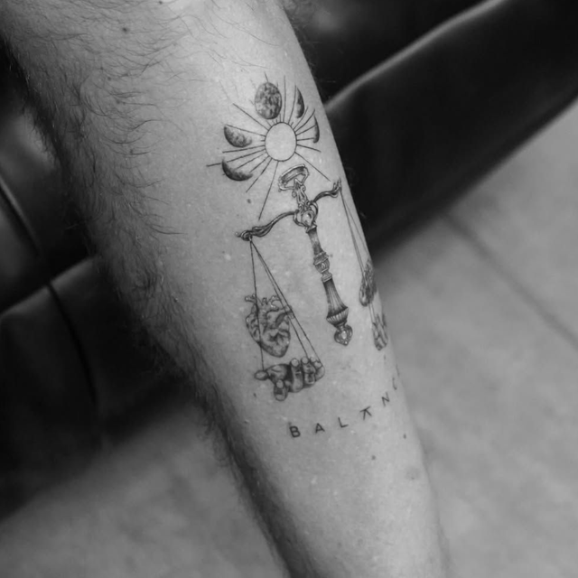 A black-ink tattoo on skin featuring a sun and moon phases above a scale weighing a heart against a brain.
