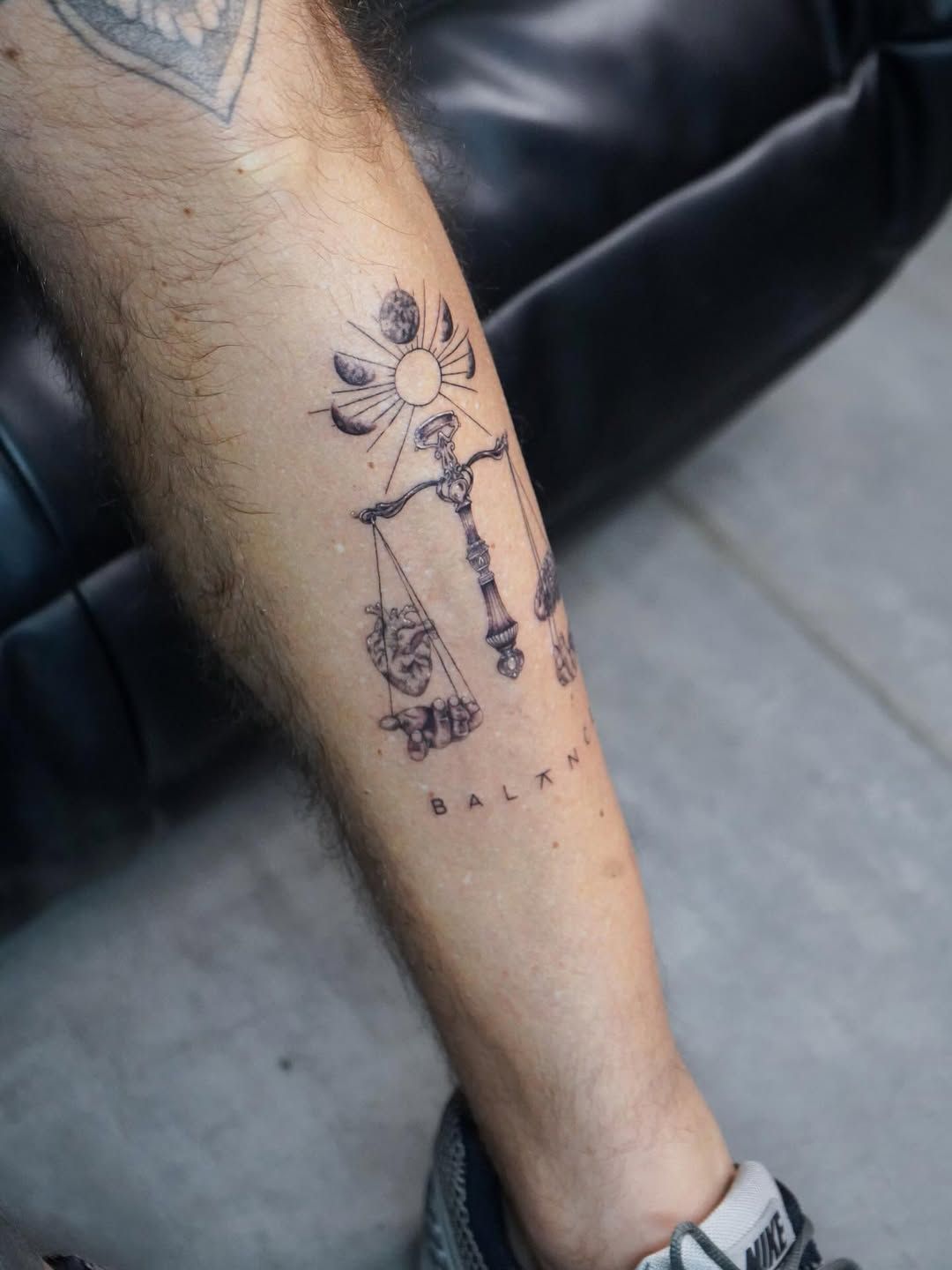 A tattoo on a forearm featuring a scale of justice balanced by a sun and lunar phases, with the word 