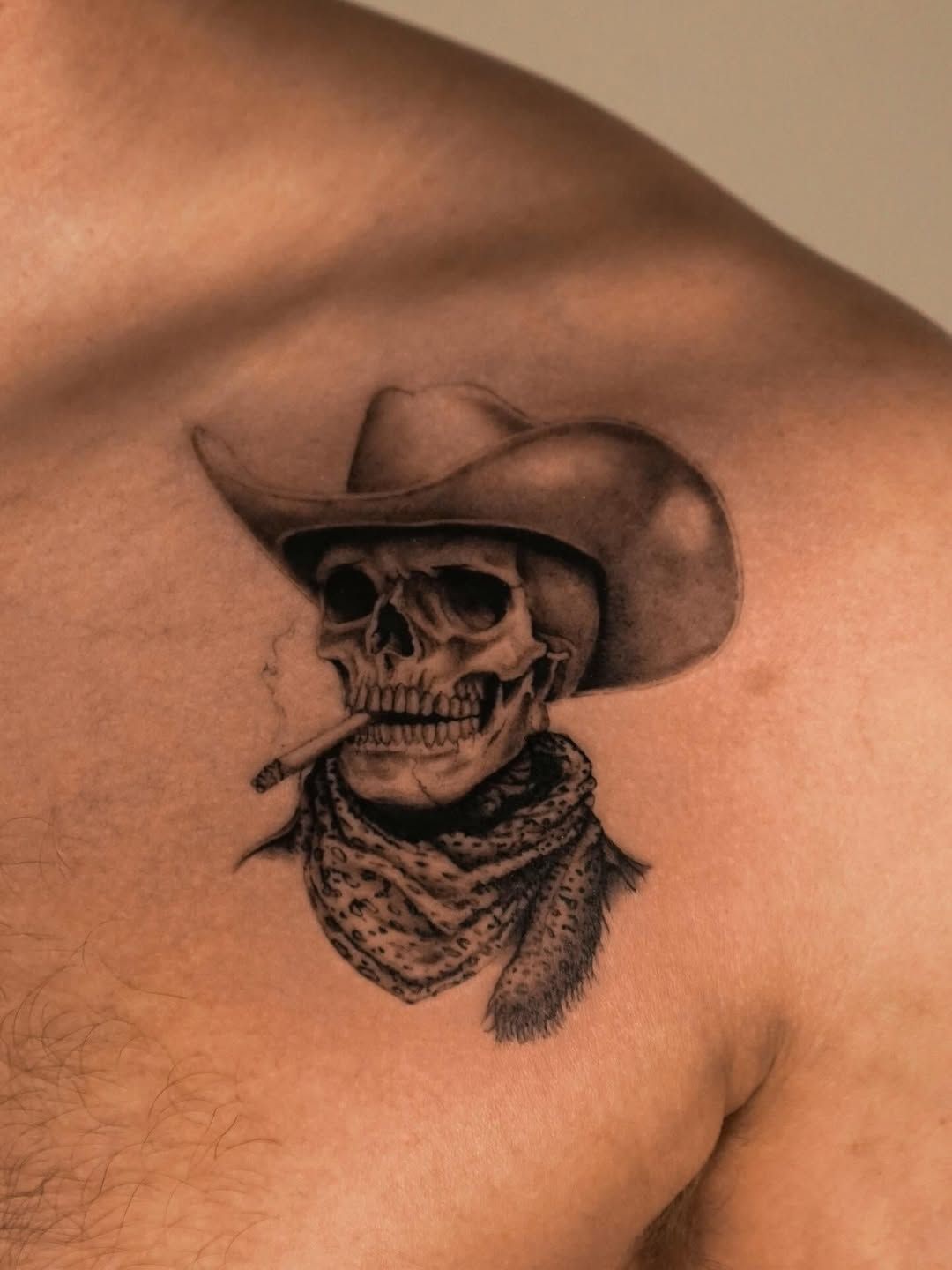 A black-and-grey tattoo on a shoulder featuring a skull wearing a cowboy hat and a bandana while smoking a cigarette.