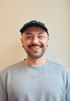 A person wearing a dark baseball cap, earrings, and a chain necklace smiles at the camera against a beige wall.