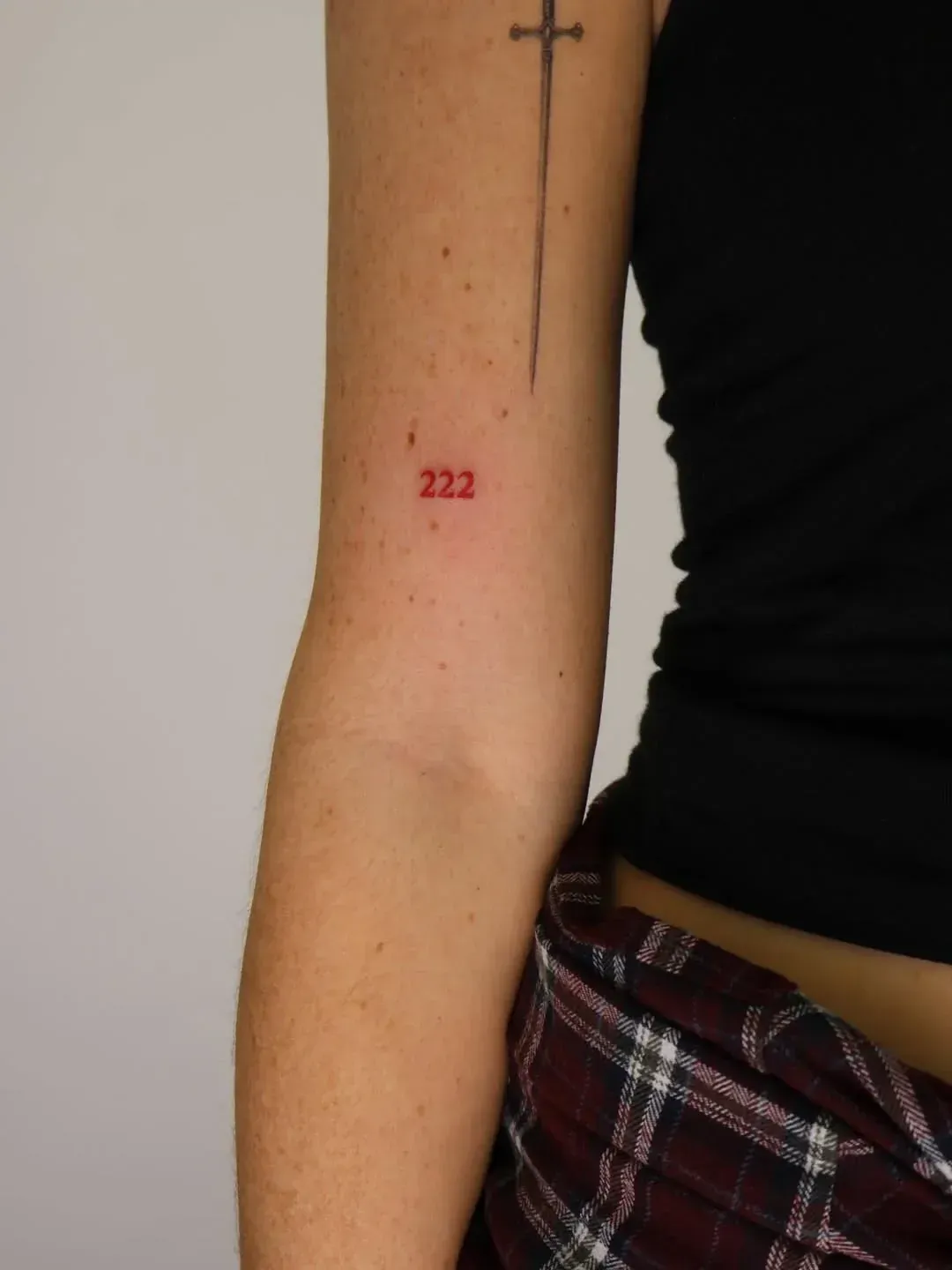 A person's upper arm showing a thin black tattoo of a sword above a small, red numerical tattoo of 