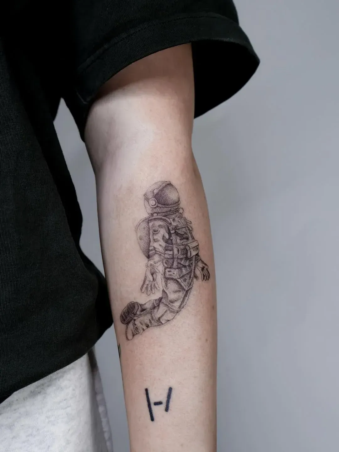 A person's forearm featuring a detailed, black-ink astronaut tattoo above a Twenty One Pilots logo tattoo.