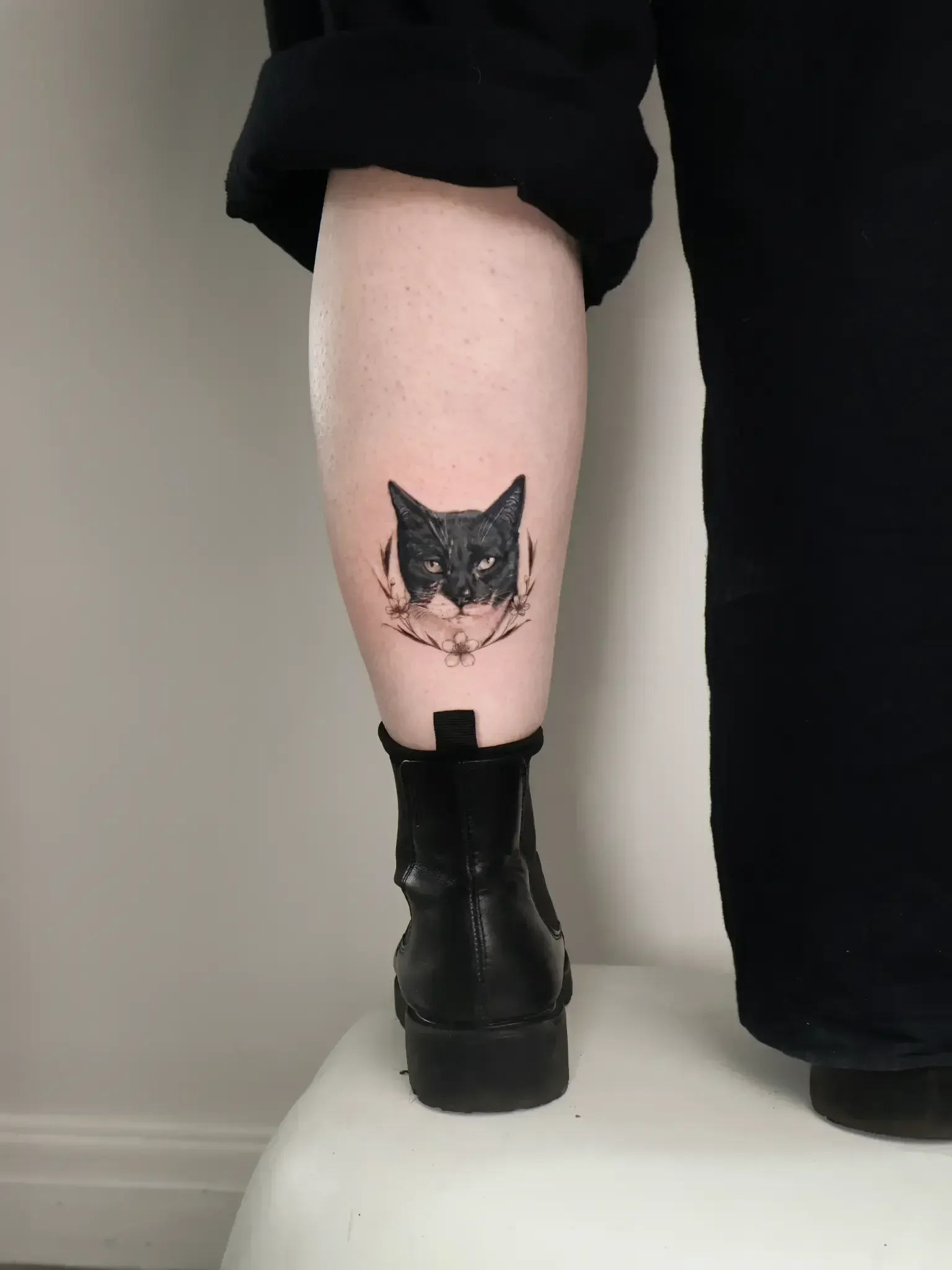 A black cat portrait tattoo with delicate floral accents positioned on the lower leg, above a black leather boot.