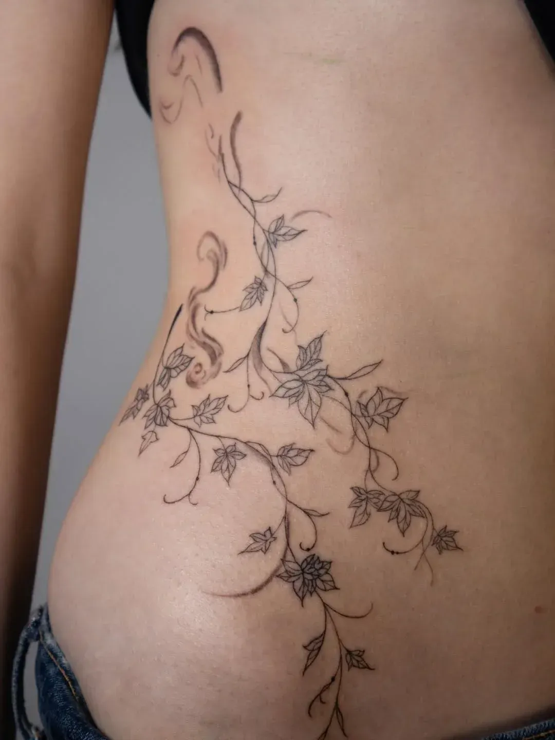 A fine-line tattoo of delicate vines with small leaves and blossoms flowing down the side of a torso.
