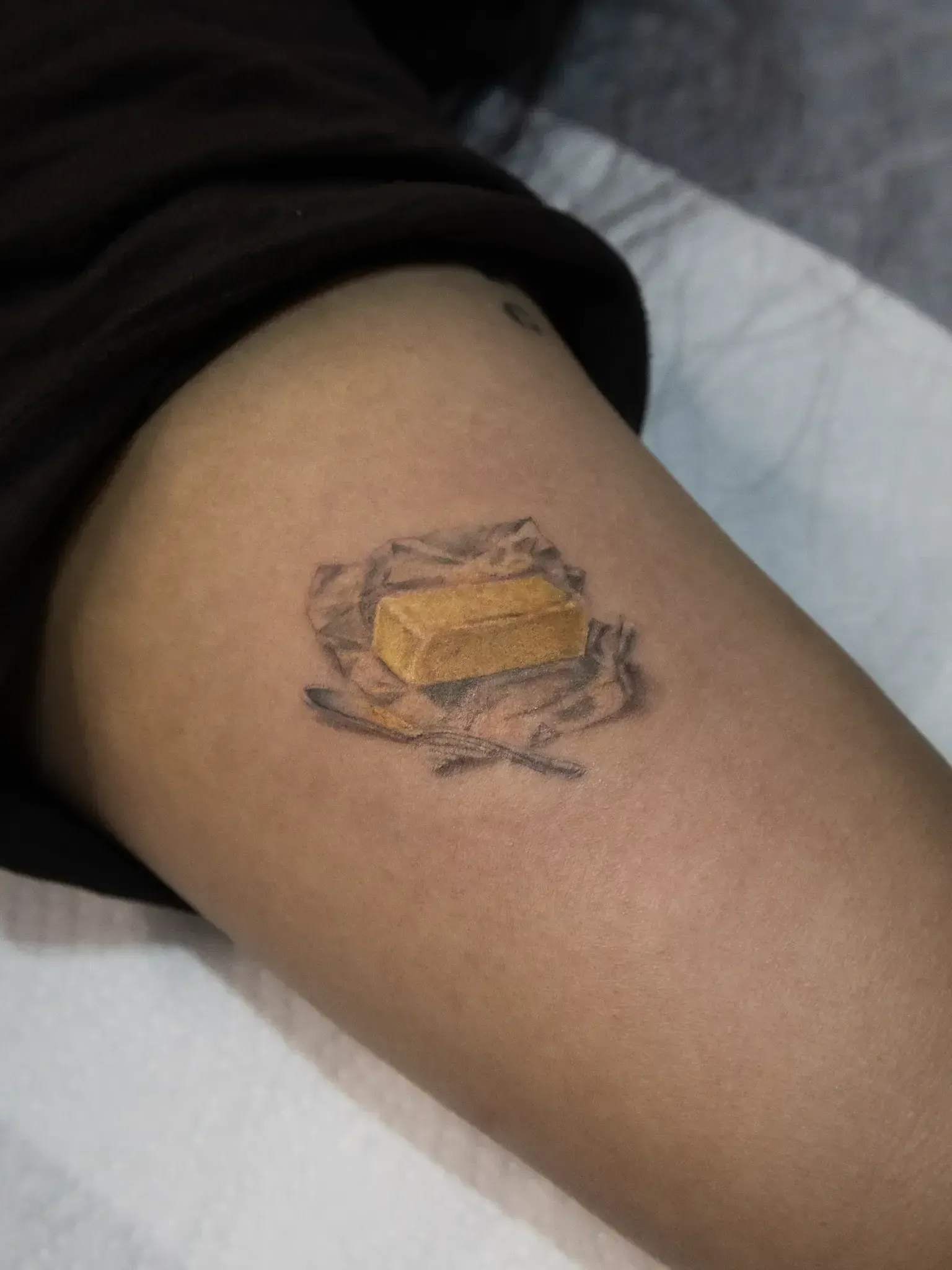 A small, realistic tattoo of a stick of yellow butter sitting on its crinkled, translucent paper wrapper.